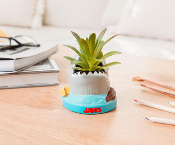 Small potted plant in a 'Jaws' themed pot on a desk with books and pencils.
